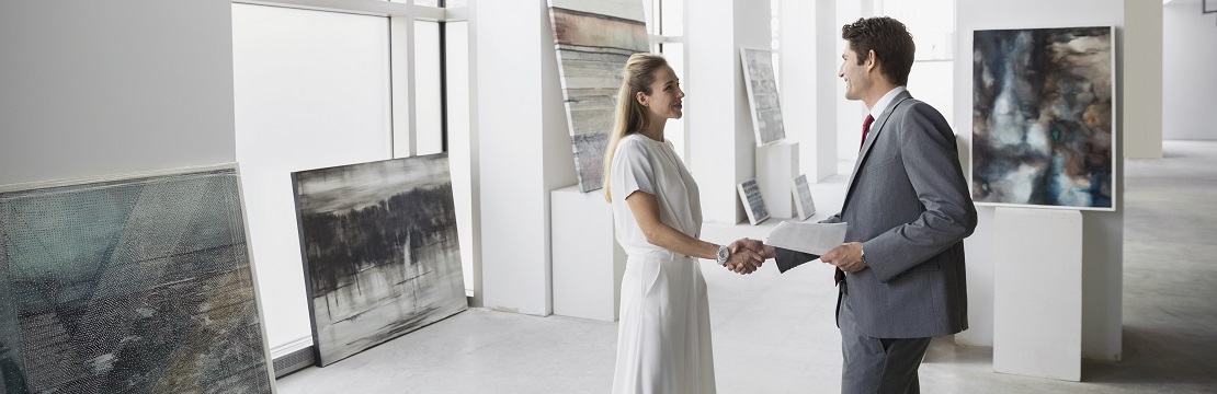 A man and woman shake hands in an art gallery with paintings leaning against the walls